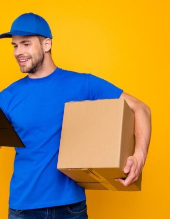 Portrait of handsome attractive cheerful bearded mailman, postman in blue uniform holding large big mail cardboard box, checking destination in clipboard, isolated over bright vivid yellow background